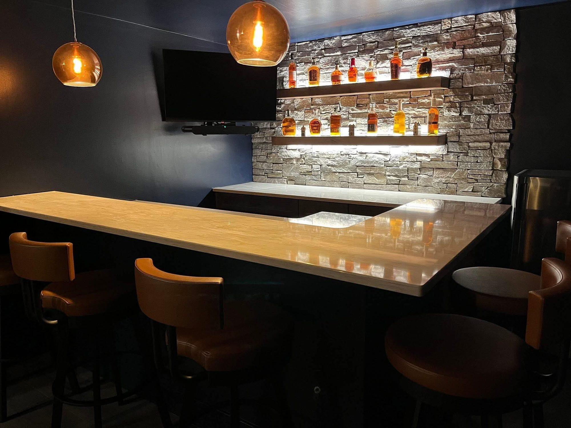 Speakeasy-style basement bar with a stacked-stone wall, lighted floating shelves, copper pendant lights and leather bar stools