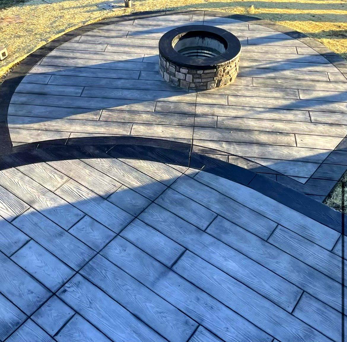 Gray wood-plank stamped concrete patio with a stone fire pit and charcoal curved accent border